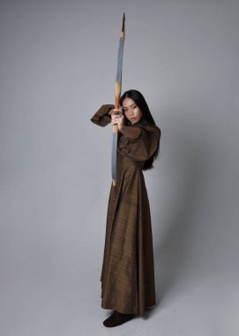 Orta çağ fantezi elbisesi giymiş, uzun saçlı güzel Asyalı bir kadının portresi. Uzun bir yay ve ok tutan zarif bir poz, stüdyo arka planında izole edilmiş..