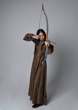 Orta çağ fantezi elbisesi giymiş, uzun saçlı güzel Asyalı bir kadının portresi. Uzun bir yay ve ok tutan zarif bir poz, stüdyo arka planında izole edilmiş..