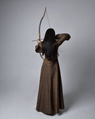 Orta çağ fantezi elbisesi giymiş, uzun saçlı güzel Asyalı bir kadının portresi. Uzun bir yay ve ok tutan zarif bir poz, stüdyo arka planında izole edilmiş..