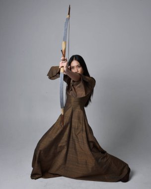 Orta çağ fantezi elbisesi giymiş, uzun saçlı güzel Asyalı bir kadının portresi. Uzun bir yay ve ok tutan zarif bir poz, stüdyo arka planında izole edilmiş..