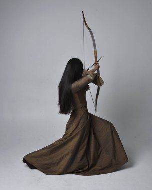 Orta çağ fantezi elbisesi giymiş, uzun saçlı güzel Asyalı bir kadının portresi. Uzun bir yay ve ok tutan zarif bir poz, stüdyo arka planında izole edilmiş..