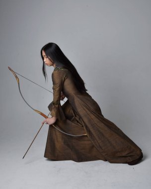 Orta çağ fantezi elbisesi giymiş, uzun saçlı güzel Asyalı bir kadının portresi. Uzun bir yay ve ok tutan zarif bir poz, stüdyo arka planında izole edilmiş..