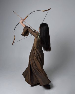 Orta çağ fantezi elbisesi giymiş, uzun saçlı güzel Asyalı bir kadının portresi. Uzun bir yay ve ok tutan zarif bir poz, stüdyo arka planında izole edilmiş..