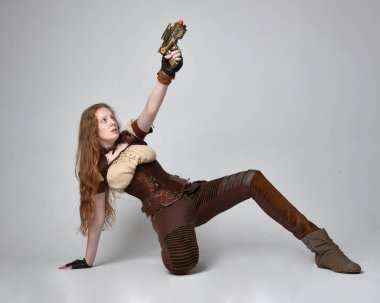Uzun kızıl saçlı, Steampunk 'tan esinlenilmiş bir kostüm giymiş güzel bir genç kadının tam boy portresi. Silah tutarken poz vermek, stüdyo arka planında izole edilmiş..