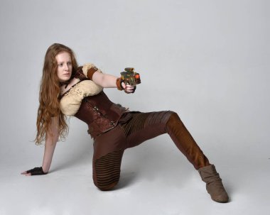 Uzun kızıl saçlı, Steampunk 'tan esinlenilmiş bir kostüm giymiş güzel bir genç kadının tam boy portresi. Silah tutarken poz vermek, stüdyo arka planında izole edilmiş..