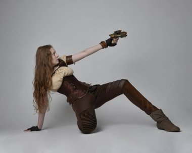 Uzun kızıl saçlı, Steampunk 'tan esinlenilmiş bir kostüm giymiş güzel bir genç kadının tam boy portresi. Silah tutarken poz vermek, stüdyo arka planında izole edilmiş..