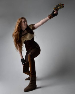 Uzun kızıl saçlı, Steampunk 'tan esinlenilmiş bir kostüm giymiş güzel bir genç kadının tam boy portresi. Silah tutarken poz vermek, stüdyo arka planında izole edilmiş..