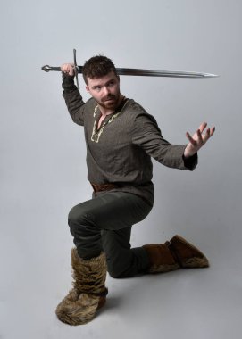 Orta Çağ Kelt maceraperest kostümü giymiş yakışıklı bir adamın tam boy portresi. Stüdyo arka planında izole edilmiş bir kılıçla ayakta duruyor..