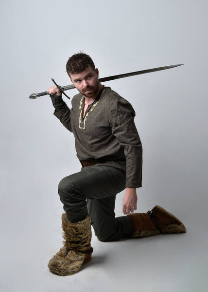 Full length  portrait of  young handsome man  wearing  medieval Celtic adventurer costume.  Standing pose holding a sword, isolated on studio background.