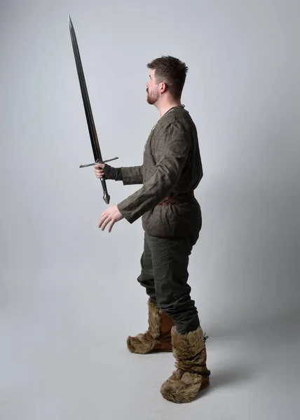 年轻英俊男子穿着中世纪凯尔特冒险家服装的全长肖像 拿着剑站在那里 在工作室的背景下与世隔绝 — 图库照片