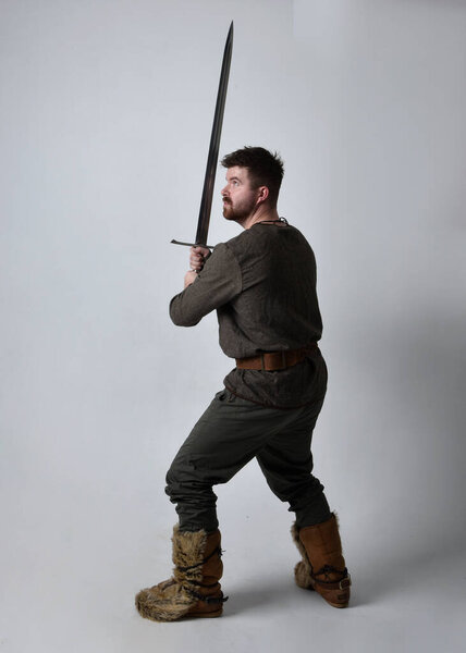 Full length  portrait of  young handsome man  wearing  medieval Celtic adventurer costume.  Standing pose holding a sword, isolated on studio background.
