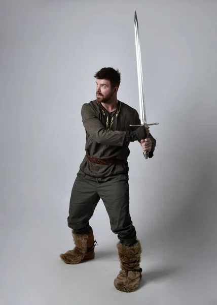 中世のケルトの冒険家の衣装を着た若いハンサムな男の完全な長さの肖像画 剣を持って立ちポーズ スタジオの背景に隔離された — ストック写真