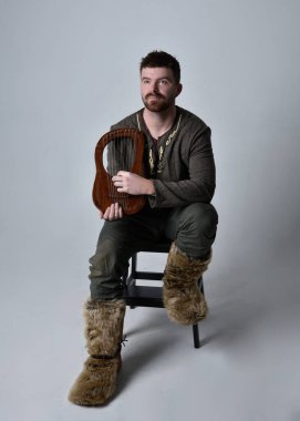 Orta çağ Kelt maceracı kostümü giymiş, stüdyo arka planında izole edilmiş küçük bir müzik arpı çalan yakışıklı bir adamın tam boy portresi..