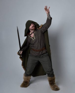 Orta çağ Kelt maceraperest kostümü giymiş kapüşonlu pelerinli, elinde kılıçla stüdyo arka planında izole edilmiş yakışıklı bir adamın tam boy portresi..