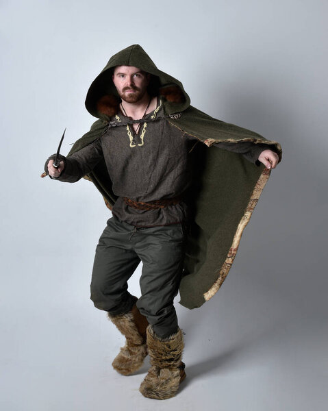 Full length  portrait of  young handsome man  wearing  medieval Celtic adventurer costume with hooded cloak, holding sword, isolated on studio background.