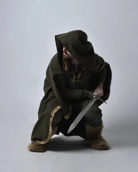 中世のケルトの冒険家の衣装を身に着けている若いハンサムな男の完全な長さの肖像画フード付きのマント 剣を保持 スタジオの背景に孤立 — ストック写真