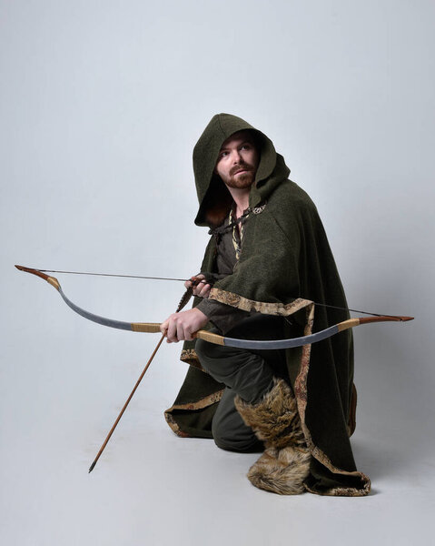 Портрет молодого красавца в средневековом костюме кельтского авантюриста в капюшоне, с луком и стрелами, изолированным на фоне студии.