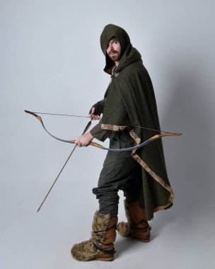 Full length  portrait of  young handsome man  wearing  medieval Celtic adventurer costume with hooded cloak, holding  archery bow and arrow, isolated on studio background. clipart