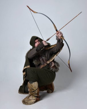 Orta çağ Kelt maceraperest kostümü giymiş kapüşonlu pelerinli, elinde okçuluk yayı ve okla stüdyo arka planında izole edilmiş genç yakışıklı bir adamın tam boy portresi..