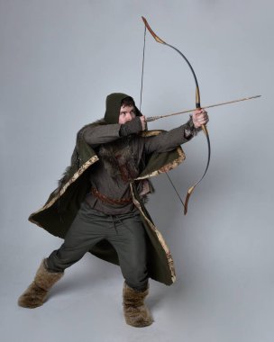 Orta çağ Kelt maceraperest kostümü giymiş kapüşonlu pelerinli, elinde okçuluk yayı ve okla stüdyo arka planında izole edilmiş genç yakışıklı bir adamın tam boy portresi..