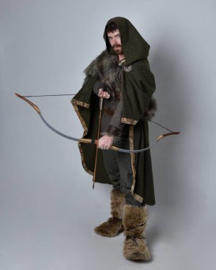 Full length  portrait of  young handsome man  wearing  medieval Celtic adventurer costume with hooded cloak, holding  archery bow and arrow, isolated on studio background. clipart
