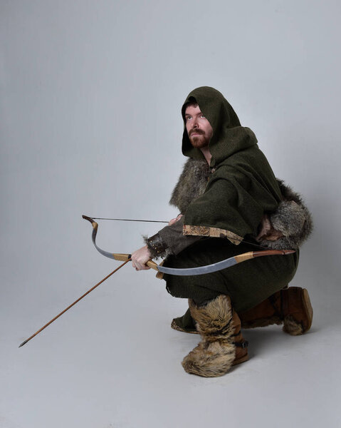 Full length  portrait of  young handsome man  wearing  medieval Celtic adventurer costume with hooded cloak, holding  archery bow and arrow, isolated on studio background.