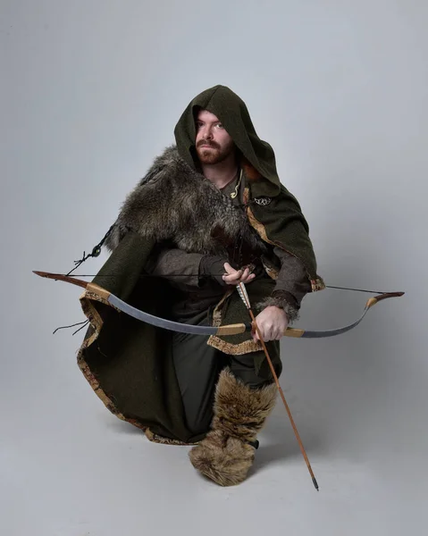 中世のケルトの冒険家の衣装を身に着けている若いハンサムな男の完全な長さの肖像画フード付きのマント 弓と矢を保持 スタジオの背景に孤立 — ストック写真