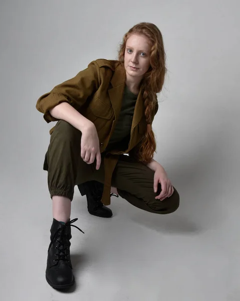 全长肖像漂亮的红头发女人穿着陆军绿色卡其布衬衫 实用的裤子和靴子 独立于工作室背景的坐姿 — 图库照片