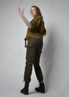 Uzun boylu, kızıl saçlı, ordu yeşili haki gömlekli, faydacı pantolon ve çizme giyen bir kadın portresi. Kameraya dönük poz, stüdyo arka planında izole edilmiş..