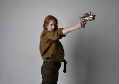 Uzun boylu, kızıl saçlı, ordu yeşili haki gömlekli, faydacı pantolon ve çizme giyen bir kadın portresi. Silah olarak silah tutmak, stüdyo arka planında izole edilmiş..