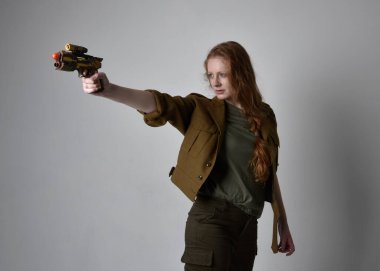 Uzun boylu, kızıl saçlı, ordu yeşili haki gömlekli, faydacı pantolon ve çizme giyen bir kadın portresi. Silah olarak silah tutmak, stüdyo arka planında izole edilmiş..