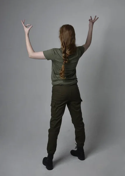 軍の緑のカーキシャツ 実用的なパンツやブーツを身に着けているかなり赤い髪の女性の完全な長さの肖像画 カメラに背を向けて立ちポーズ スタジオの背景に隔離されました — ストック写真