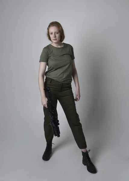 Full length portrait of pretty red haired woman wearing army green khaki shirt, utilitarian pants and boots. Holding a gun as weapon, isolated on studio background.