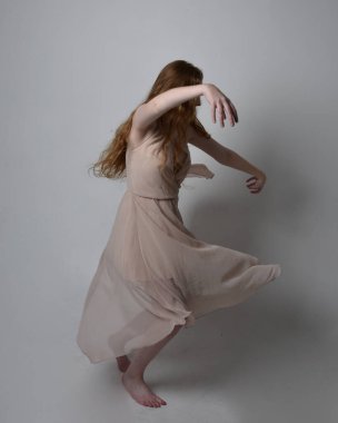 Güzel kızıl saçlı bir dansçının uzun boylu portresi. Ten rengi, akıcı peri elbisesi giyiyor. Ayakta duran hareketli pozlar stüdyo arka planında izole.