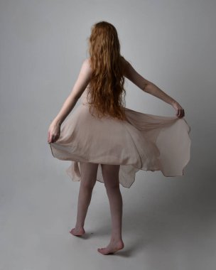 Güzel kızıl saçlı bir dansçının uzun boylu portresi. Ten rengi, akıcı peri elbisesi giyiyor. Ayakta duran hareketli pozlar stüdyo arka planında izole.