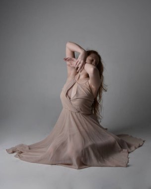 Güzel kızıl saçlı bir dansçının uzun boylu portresi. Ten rengi, akıcı peri elbisesi giyiyor. Oturan hareketli pozlar stüdyo arka planında izole.