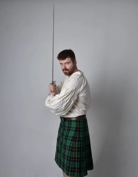Close up portrait of handsome brunette man wearing Scottish kilt and renaissance white  pirate blouse shirt. Holding a sword weapon, action pose isolated against studio background.