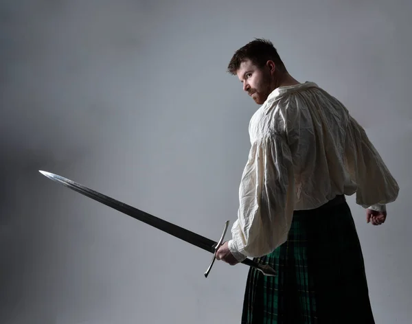 Close up portrait of handsome brunette man wearing Scottish kilt and renaissance white  pirate blouse shirt. Holding a sword weapon, action pose isolated against studio background.