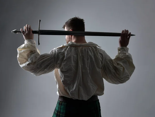 Close up portrait of handsome brunette man wearing Scottish kilt and renaissance white  pirate blouse shirt. Holding a sword weapon, action pose isolated against studio background.