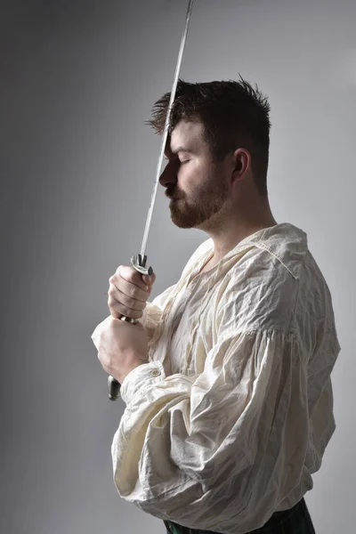 Close up portrait of handsome brunette man wearing Scottish kilt and renaissance white  pirate blouse shirt. Holding a sword weapon, action pose isolated against studio background.