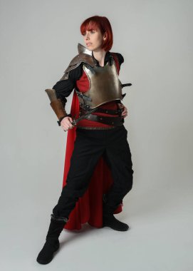 Savaşçı kadın şövalyenin tam boy fantezi portresi, tarihi ortaçağ çelik zırh kostümü giyen bir model, kırmızı peri masalı pelerini, kraliyet tacı. Diz çöküp bıçak tutan, izole edilmiş stüdyo geçmişi olan.