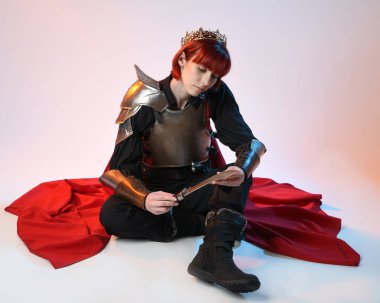 Savaşçı kadın şövalyenin tam boy fantezi portresi, tarihi ortaçağ çelik zırh kostümü giyen bir model, kırmızı peri masalı pelerini, kraliyet tacı. Diz çöküp bıçak tutan, izole edilmiş stüdyo geçmişi olan.