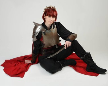 Savaşçı kadın şövalyenin tam boy fantezi portresi, tarihi ortaçağ çelik zırh kostümü giyen bir model, kırmızı peri masalı pelerini, kraliyet tacı. Diz çöküp bıçak tutan, izole edilmiş stüdyo geçmişi olan.