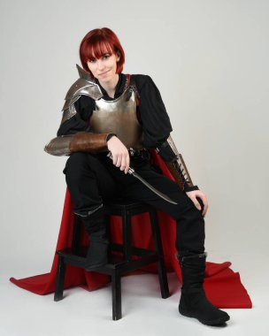 Savaşçı kadın şövalyenin fantezi portresi, tarihi ortaçağ zırhı giymiş bir model, kırmızı peri masalı pelerini. Oturma pozu bıçak hançeri, izole stüdyo arka planı.