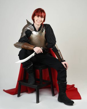 Savaşçı kadın şövalyenin fantezi portresi, tarihi ortaçağ zırhı giymiş bir model, kırmızı peri masalı pelerini. Oturma pozu bıçak hançeri, izole stüdyo arka planı.