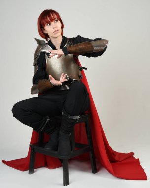 Fantezi savaşçısı kadın modelinin portresi, elinde kılıç, kırmızı peri masalı pelerinli tarihi ortaçağ zırhı, güçlü bir fantezi şövalyesi. Stüdyo arka planında izole edilmiş sihirli el hareketleriyle oturuyor.