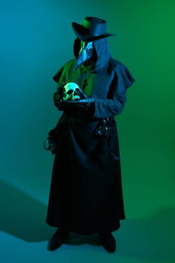Gizemli Veba Doktoru Cadılar Bayramı Portresi, Korkunç Gotik Karakter Tutucu Kafatası Yadigarı Kara Pelerinli Karanlık Stüdyo Sahnesi, Kuş Maskesi. Atmosferik Işıklı Korku Fantezi Kostümü
