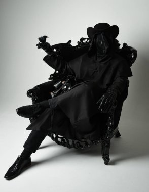 Fenerli Gotik Veba Doktoru, Kara Pelerinli Cadılar Bayramı Karakteri, Kuş Maskeli, kadife taht koltuğunda oturan. Gizemli Fantezi Korku figürünün Stüdyo Portresi