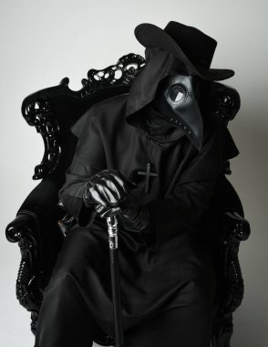 Fenerli Gotik Veba Doktoru, Kara Pelerinli Cadılar Bayramı Karakteri, Kuş Maskeli, kadife taht koltuğunda oturan. Gizemli Fantezi Korku figürünün Stüdyo Portresi