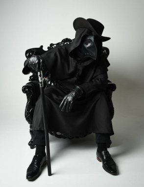 Fenerli Gotik Veba Doktoru, Kara Pelerinli Cadılar Bayramı Karakteri, Kuş Maskeli, kadife taht koltuğunda oturan. Gizemli Fantezi Korku figürünün Stüdyo Portresi